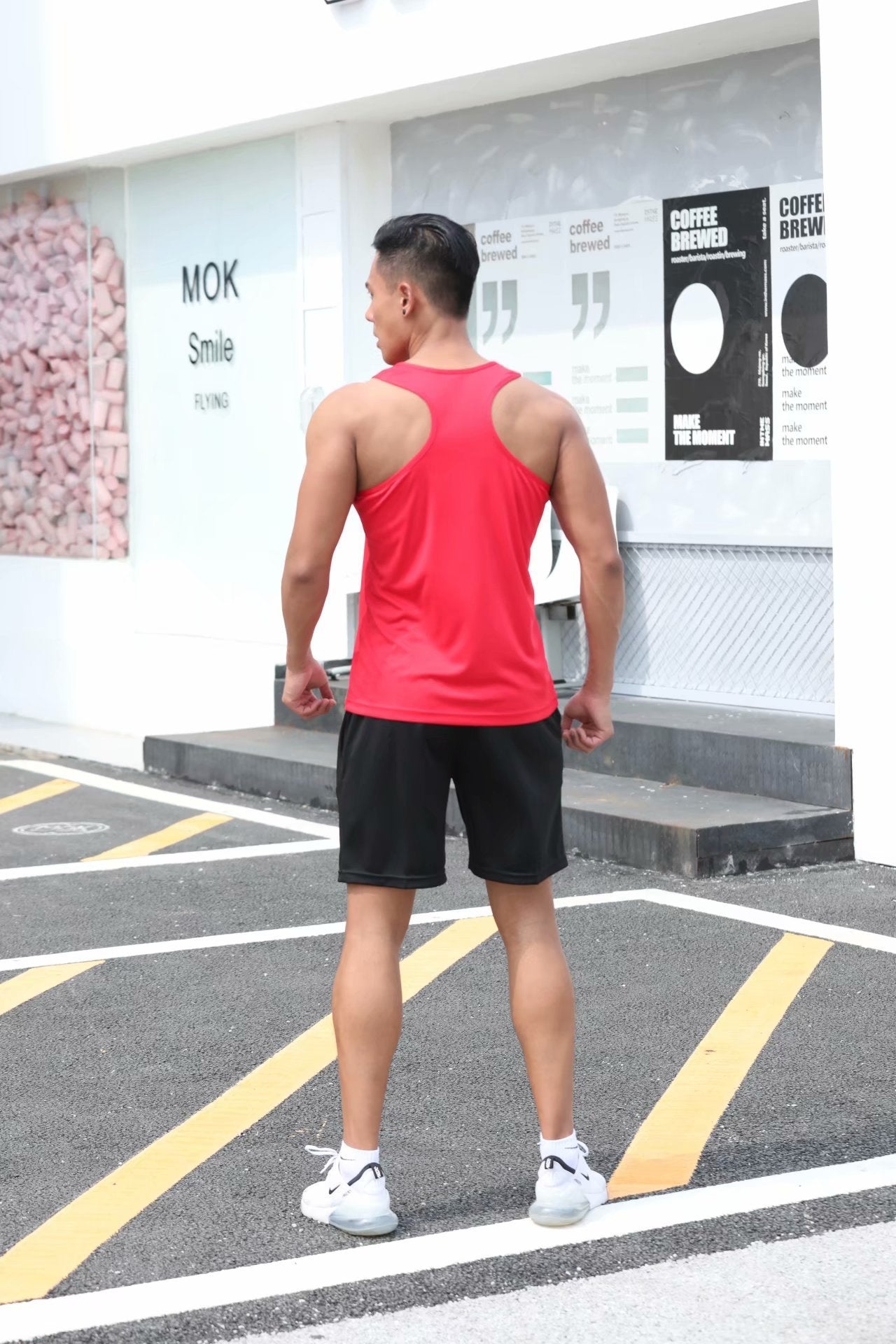 Person wearing a red tank top and black shorts standing on a street with a building in the background.