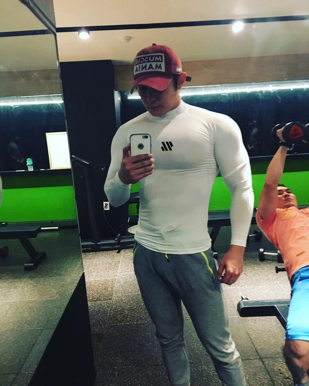 Person taking a mirror selfie in a gym wearing a white long-sleeve shirt and gray pants.