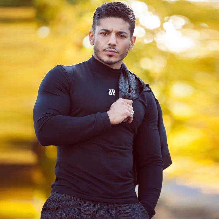Man wearing a black turtleneck with a brand logo, standing outdoors with blurred yellow foliage in the background.